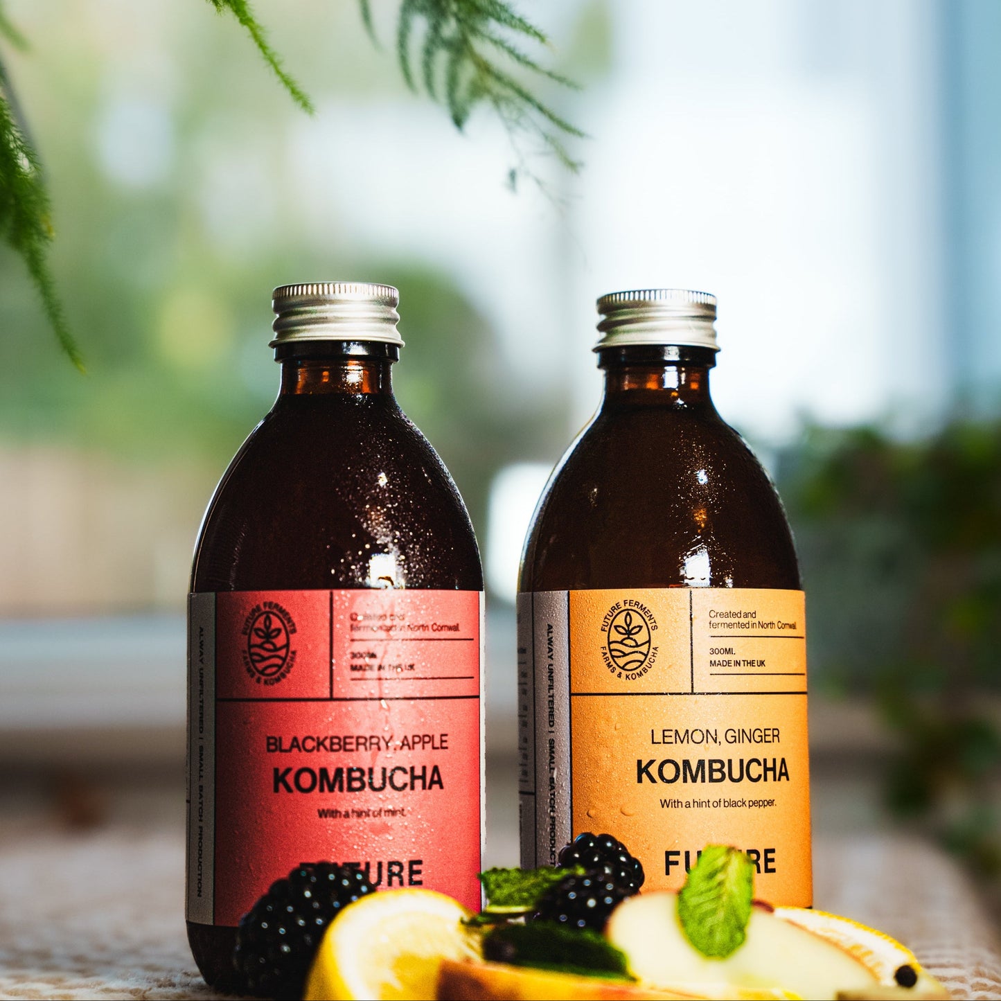 Two bottles of FUTURE Kombucha on a checkered tablecloth with blurred greenery in the background. Showing the two flavours of the mixed case of kombucha featuring Lemon Ginger Blackpepper and Blackberry Apple Mint flavours