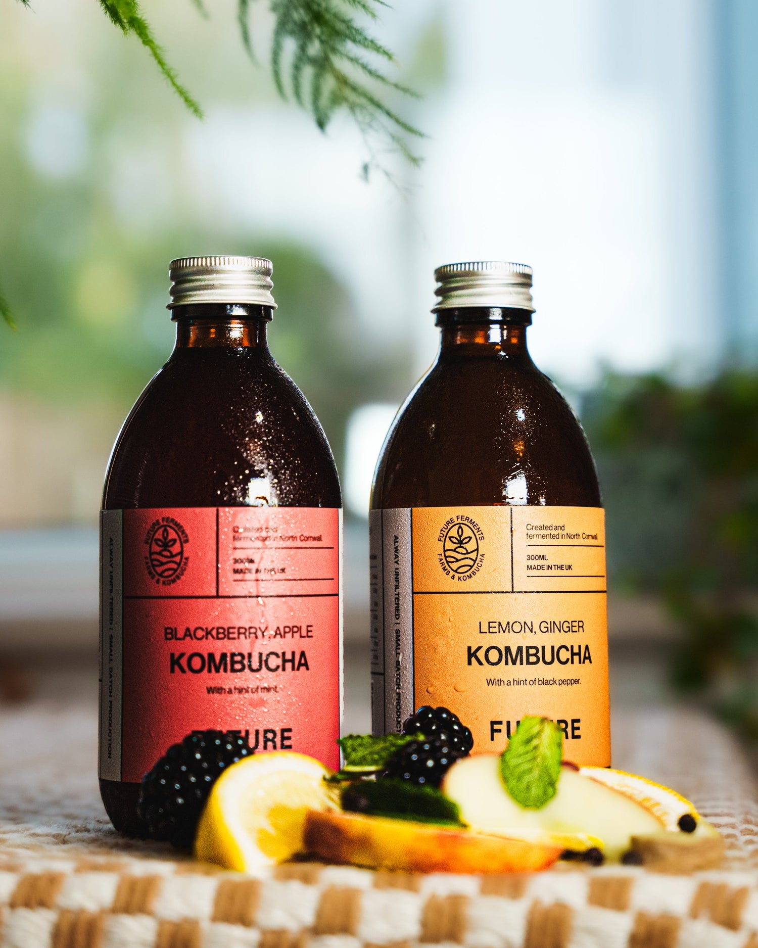 Two bottles of FUTURE Kombucha on a checkered tablecloth with blurred greenery in the background.